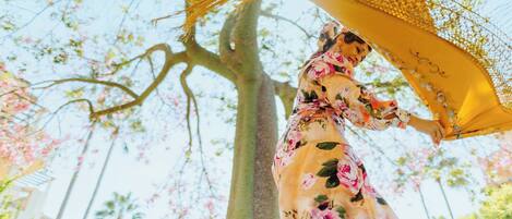 Eine Person in einem Blumenkleid hält einen gelben Sonnenschirm in einer von Bäumen gesäumten Gegend.