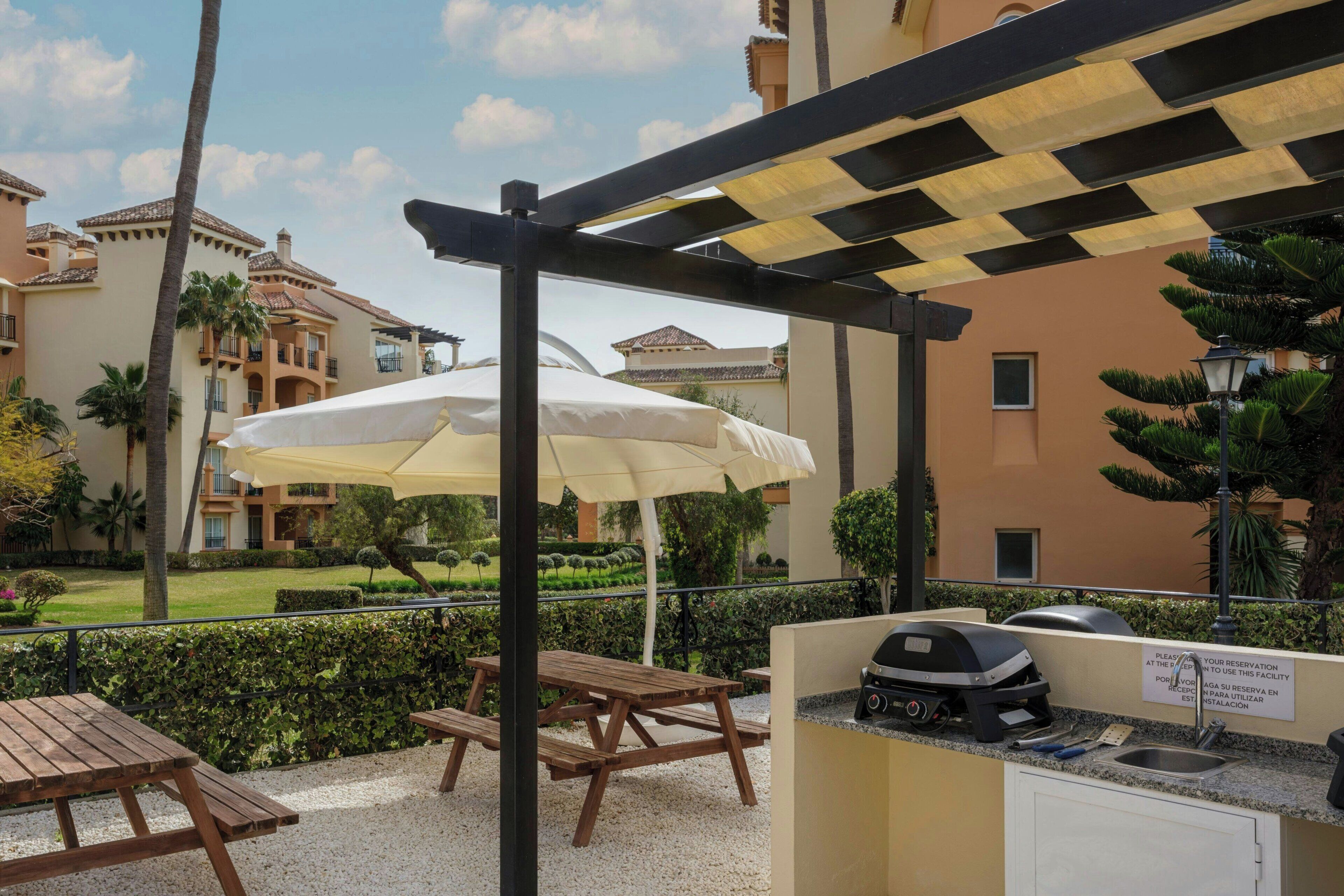 Un patio al aire libre con una parrilla, mesas de picnic y sombrillas.