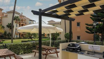 Un patio al aire libre con una parrilla, mesas de picnic y sombrillas.