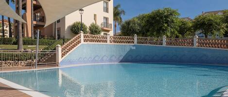 A swimming pool with a tiled edge, a palm tree, and a building with balconies in the background.