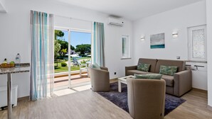 A modern living room with a sofa, armchairs, and a coffee table. There is a sliding glass door leading to an outdoor area with a pool and greenery. - Prado Villas (Loulé)