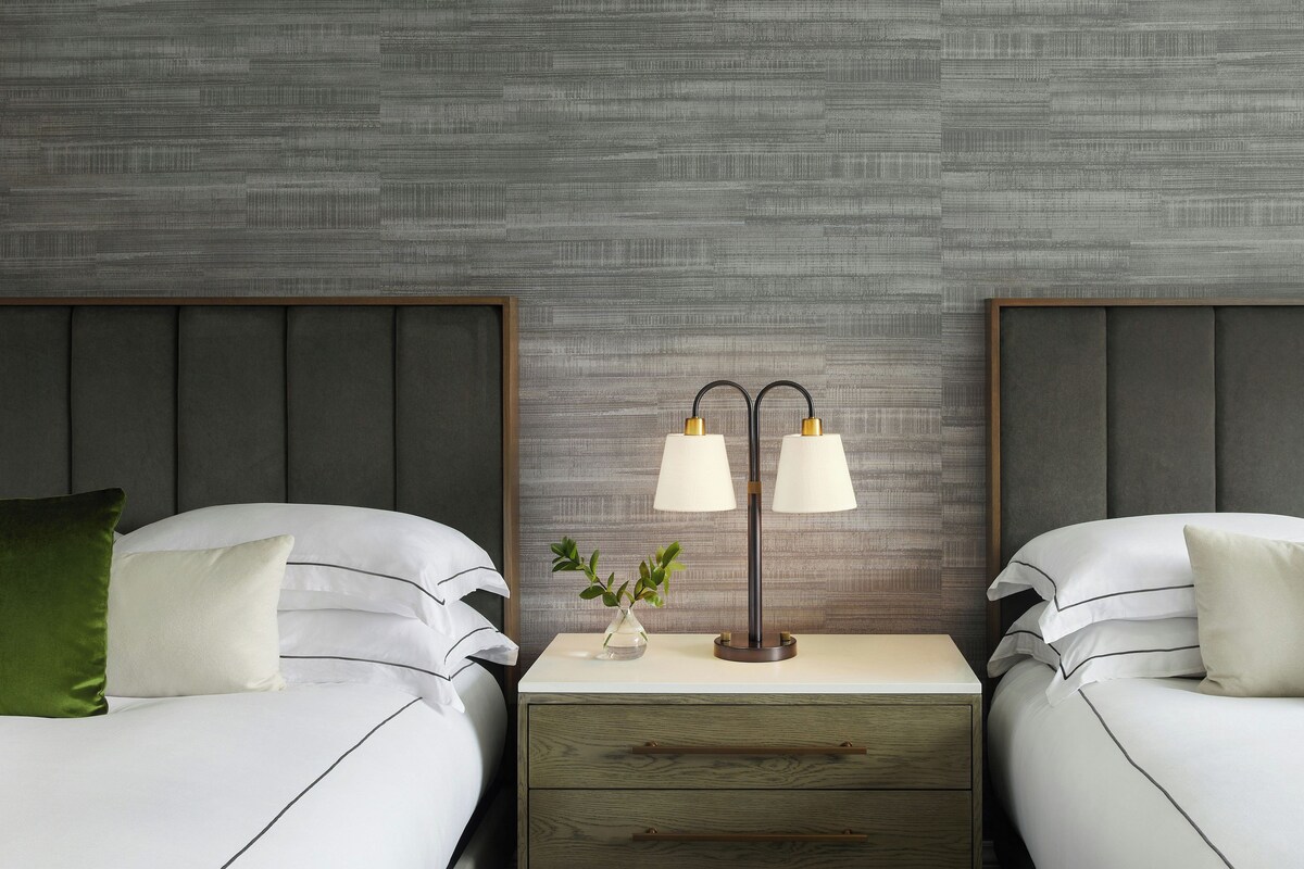 A hotel room with two beds, a nightstand with a lamp, and a textured wall.