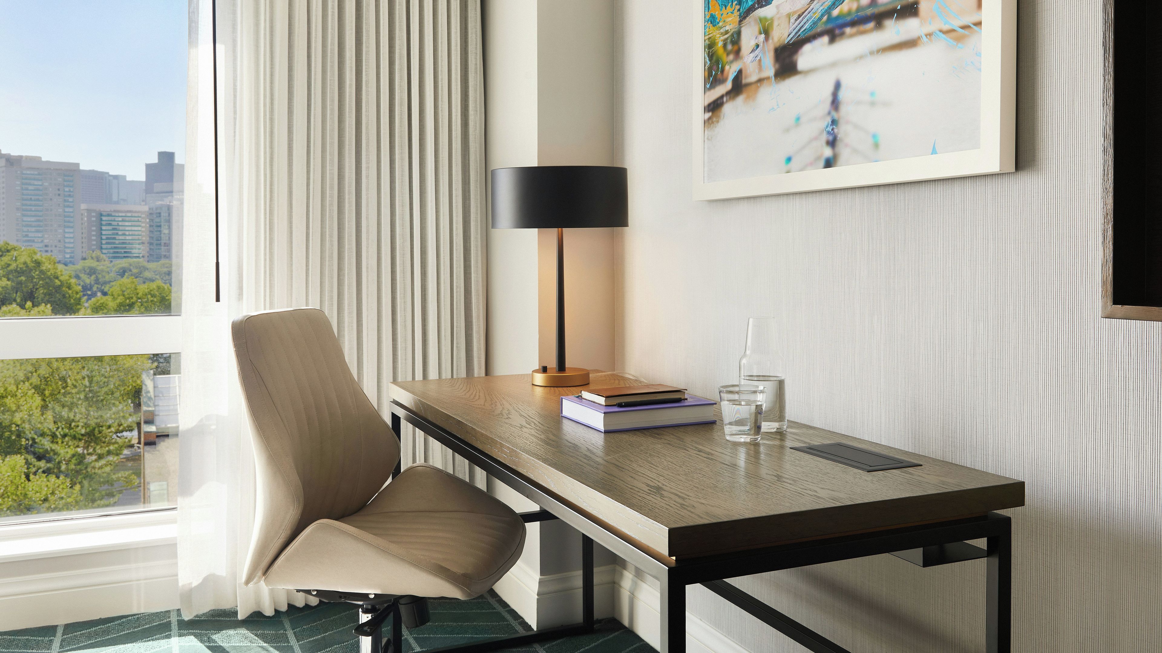 Ein Hotelzimmer mit Blick auf eine Stadtlandschaft, ein Schreibtisch mit Lampe, ein Sessel, ein Buch und ein Glas Wasser.