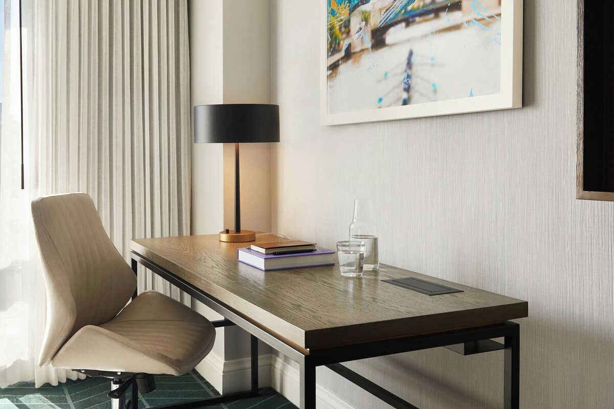 A hotel room with a view of a cityscape, a desk with a lamp, a chair, a book, and a glass of water.