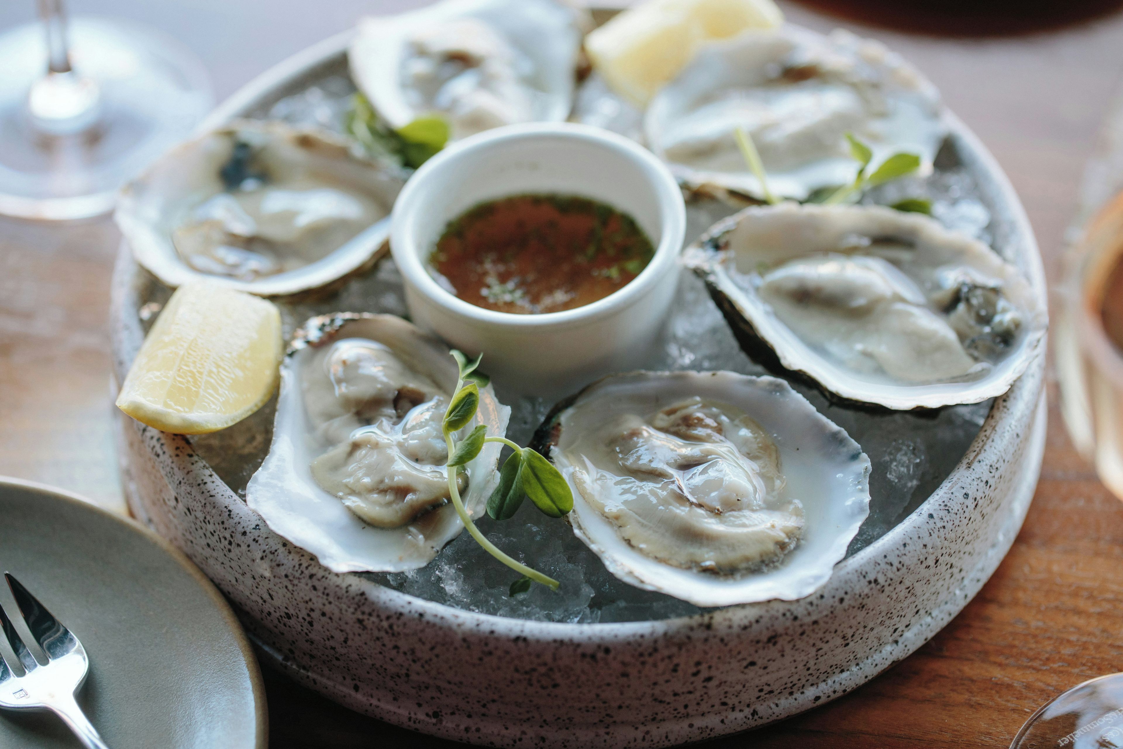 A plate of oysters on ice with lemon wedges and a small bowl of sauce.
