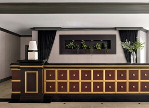 A reception desk with a dark wood finish, a black and gold decorative panel, and a plant display. - Kimpton Marlowe Hotel by IHG (Cambridge)