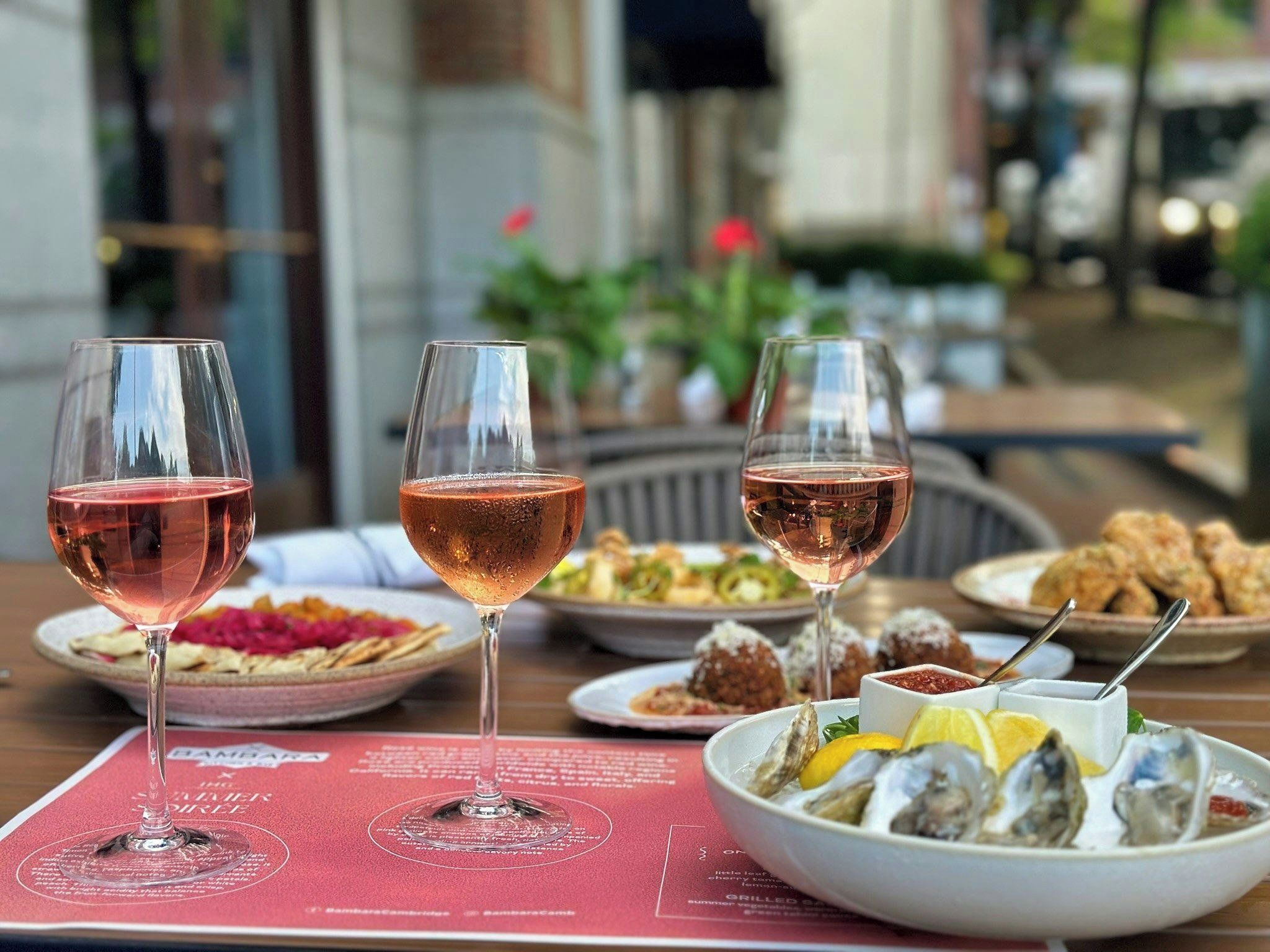 A table with three glasses of rosé wine, plates of food, and a menu.