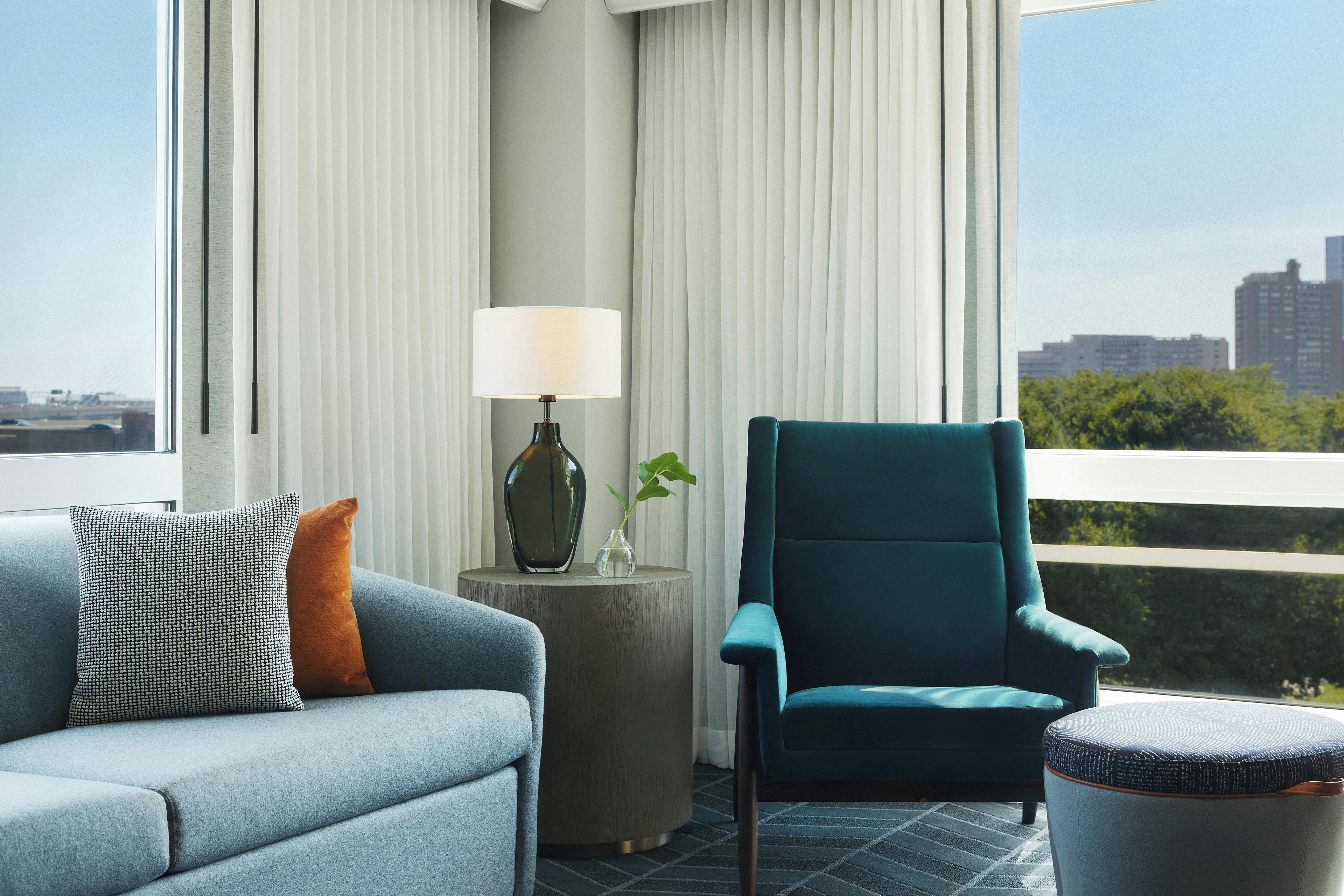 A modern living room with a sofa, armchair, and ottoman, a side table with a lamp, and a view of a cityscape through large windows.