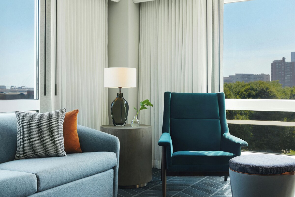 A modern living room with a sofa, armchair, and ottoman, a side table with a lamp, and a view of a cityscape through large windows.