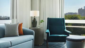 A modern living room with a sofa, armchair, and ottoman, a side table with a lamp, and a view of a cityscape through large windows.