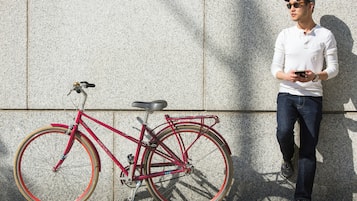 Une personne se tient à côté d’un vélo rouge, muni d’une chaîne et de pignons, et s’appuie contre un mur à la texture particulière.