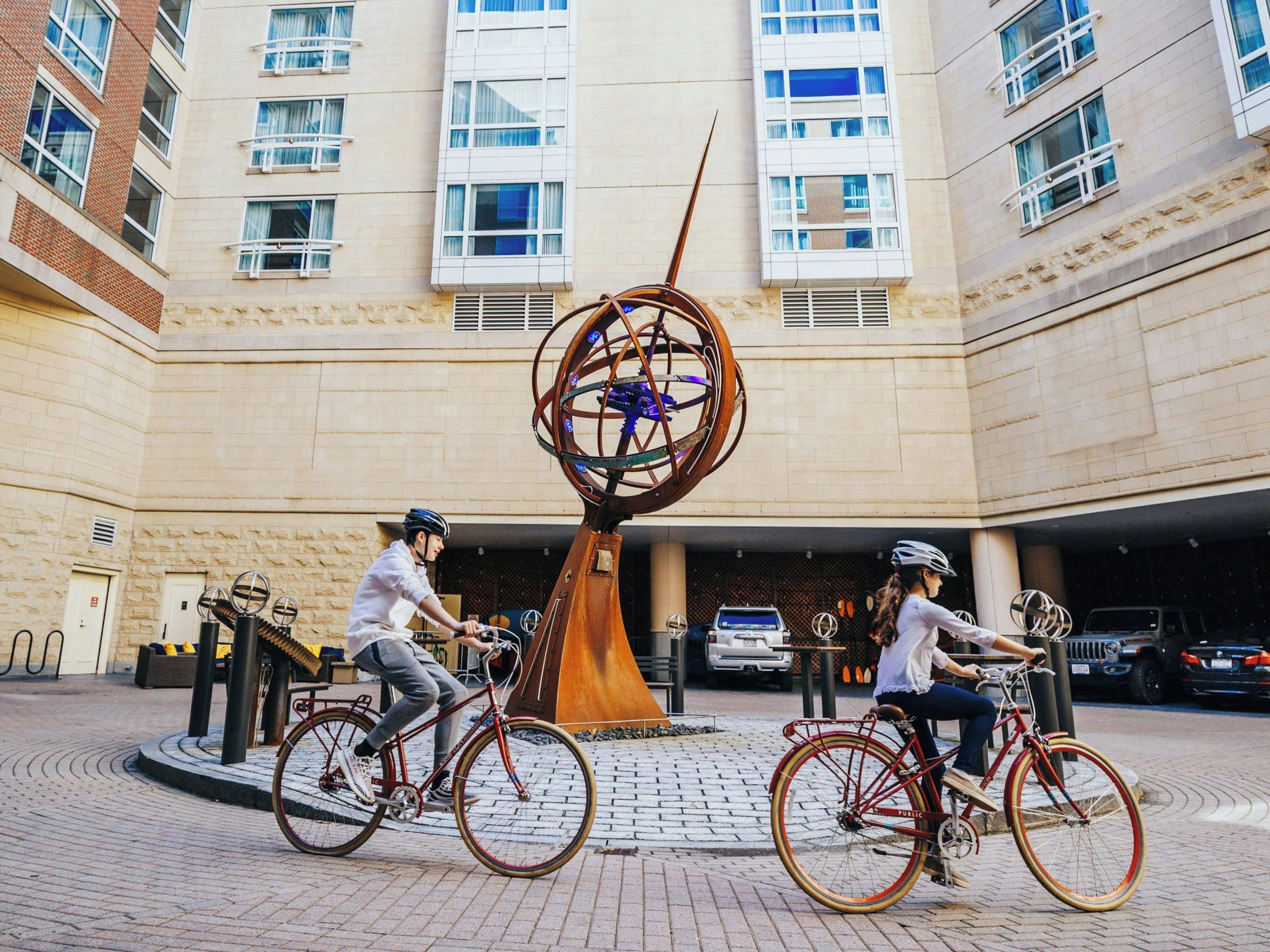 A building with a central sculpture, two cyclists, and a parking garage in the background.