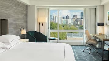 A hotel room with a large bed, two chairs, a desk, and a view of a cityscape through a large window.