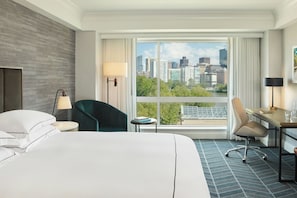 A hotel room with a large bed, two chairs, a desk, and a view of a cityscape through a large window. - Kimpton Marlowe Hotel by IHG (Cambridge)
