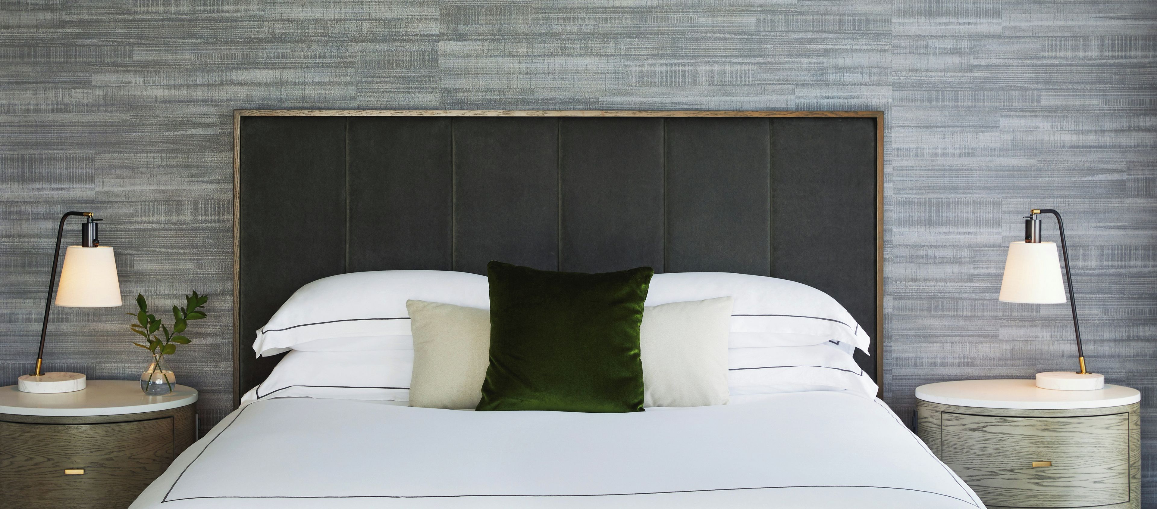 A neatly made bed with white linens and a dark headboard, flanked by two bedside tables with lamps, against a textured wall.