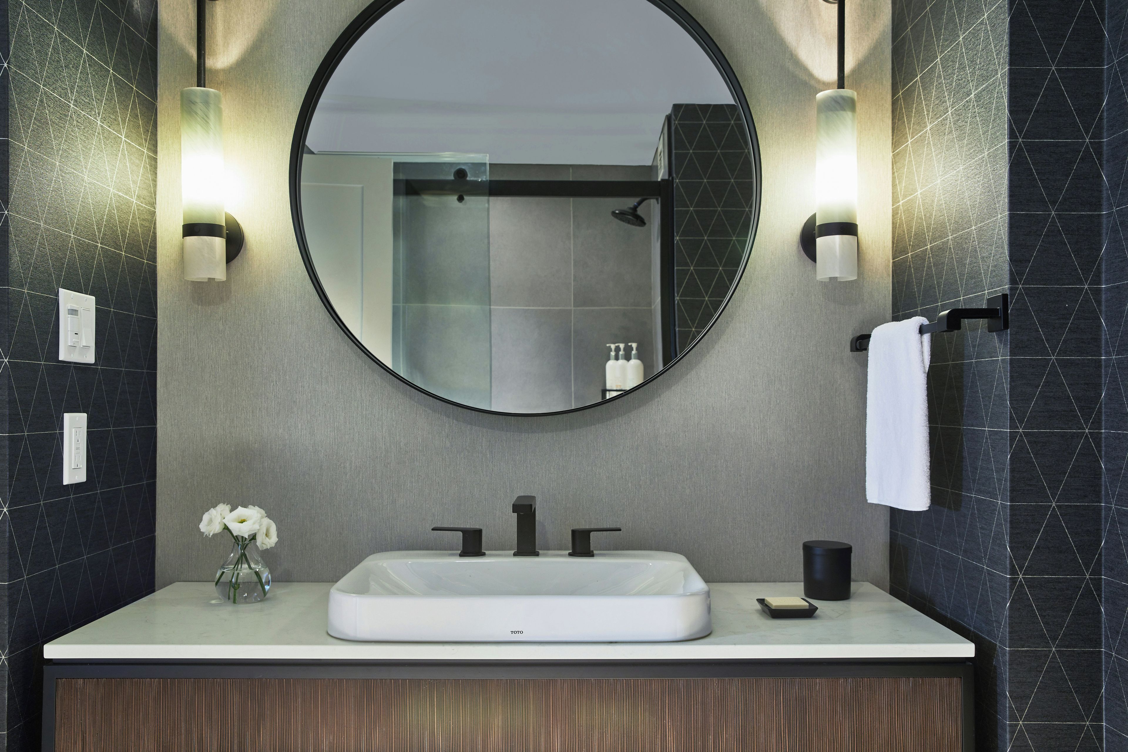 Une salle de bain moderne avec un miroir rond, un lavabo blanc et deux appliques murales.