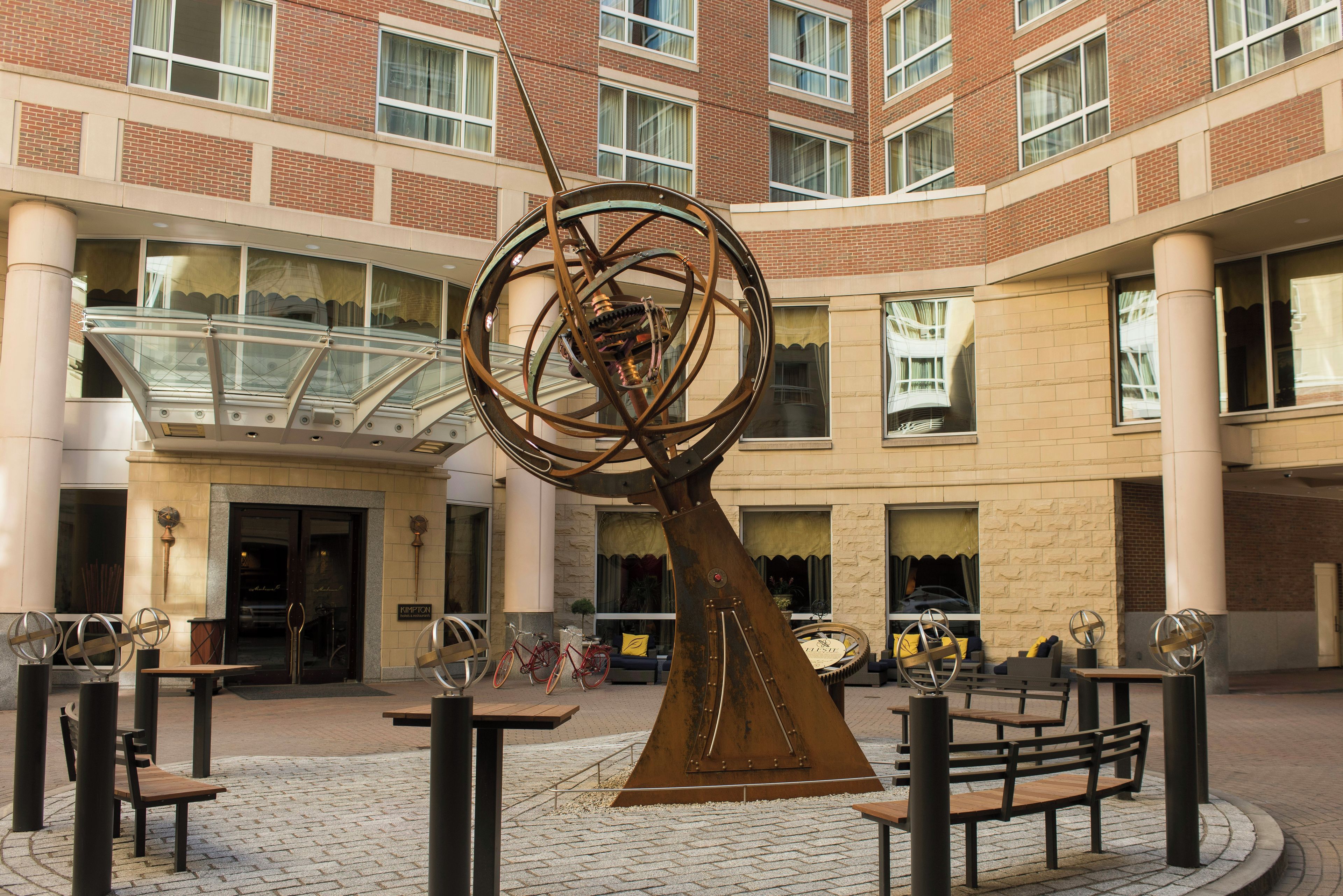 A courtyard with a large metal sculpture, seating areas, and a building with multiple windows and a glass atrium.