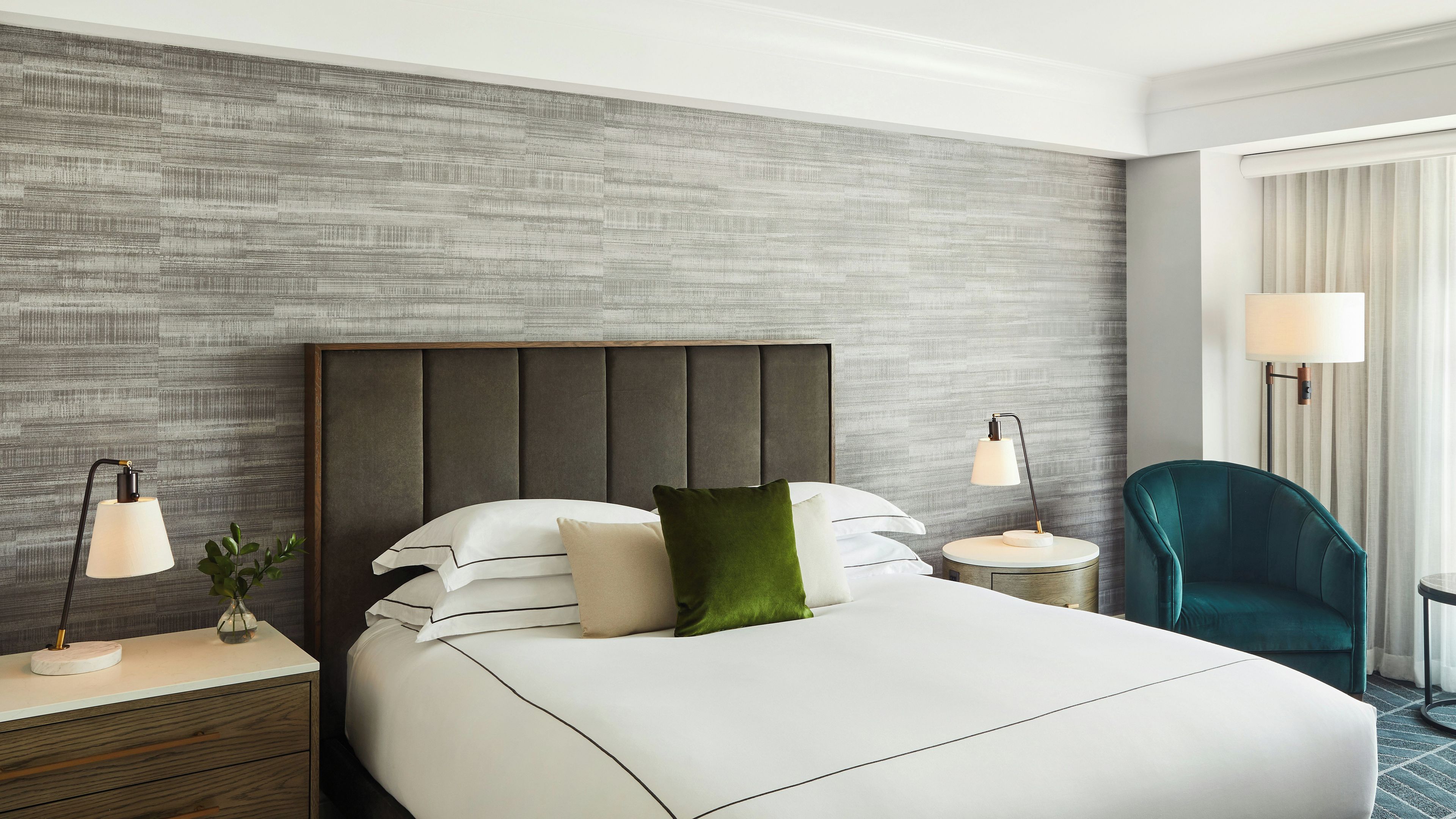 A hotel room with a large bed, two bedside tables, a green chair, and a window with curtains.