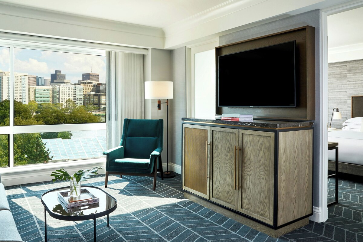 Una habitación de hotel con un ventanal grande, un sillón color aguamarina, un televisor y una cama.