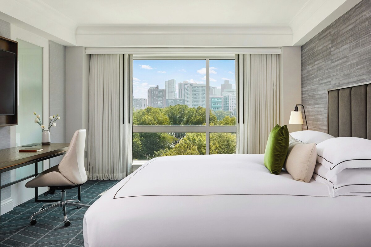 A hotel room with a large bed, a desk, a chair, a TV, and a view of the cityscape.