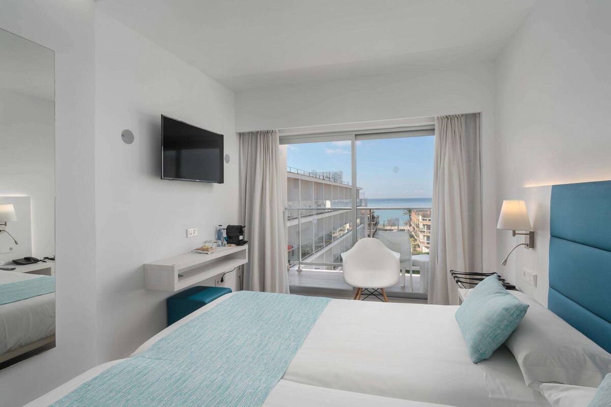 A hotel room with a bed, a desk, a chair, a TV, and a balcony with a view of the sea.