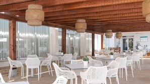 An outdoor dining area with white wicker chairs and tables, wooden beams overhead, and large windows providing natural light.