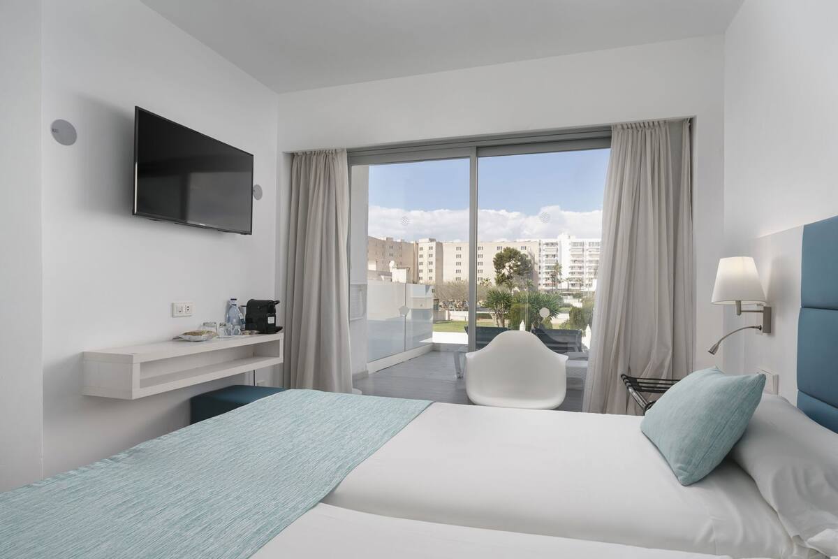A modern hotel room with a bed, a TV, a desk with a coffee maker, a chair, and a view of buildings and trees.