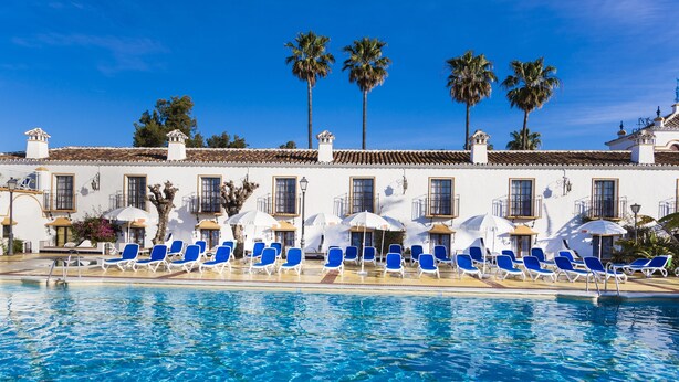 Una piscina con sillas reclinables azules y sombrillas, palmeras y un edificio blanco con varias ventanas.