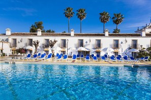 A pool with blue lounge chairs and umbrellas, palm trees, and a white building with multiple windows. - Globales Cortijo Blanco (Marbella)
