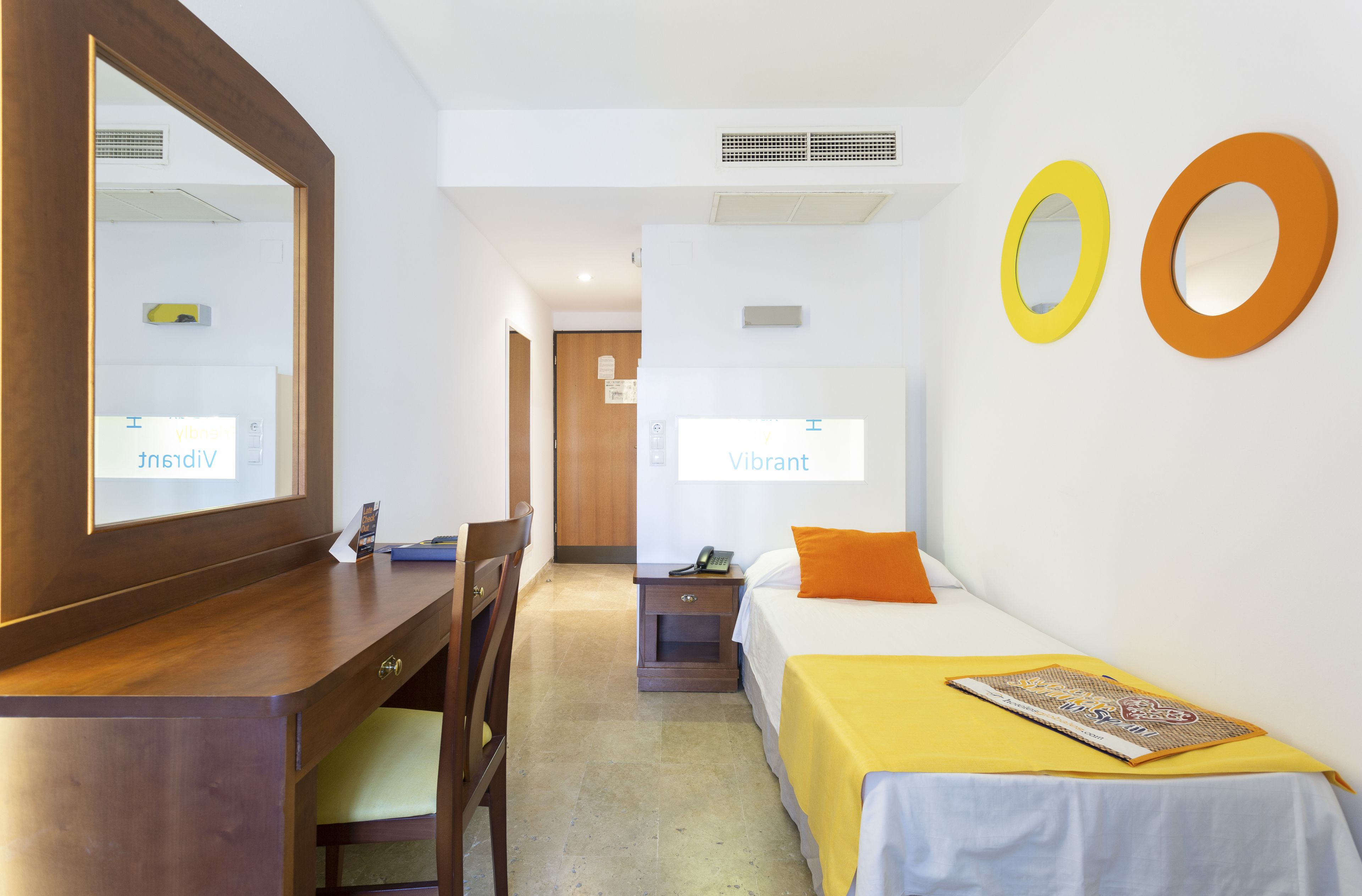 A hotel room with a wooden desk, a bed with a yellow bedspread, and two wall-mounted mirrors.