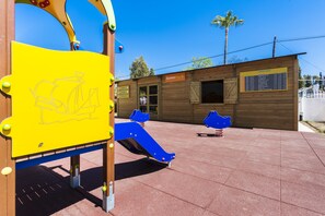 Ein Spielplatz mit einer gelben Kletterkonstruktion, einer blauen Rutsche und einem hölzernen Spielhäuschen mit dem Schriftzug „Suneo Play“.