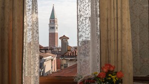 Vista a través de una ventana con cortinas, mostrando un edificio con una torre alta y una cúpula, además de un ramo de flores en el alféizar.