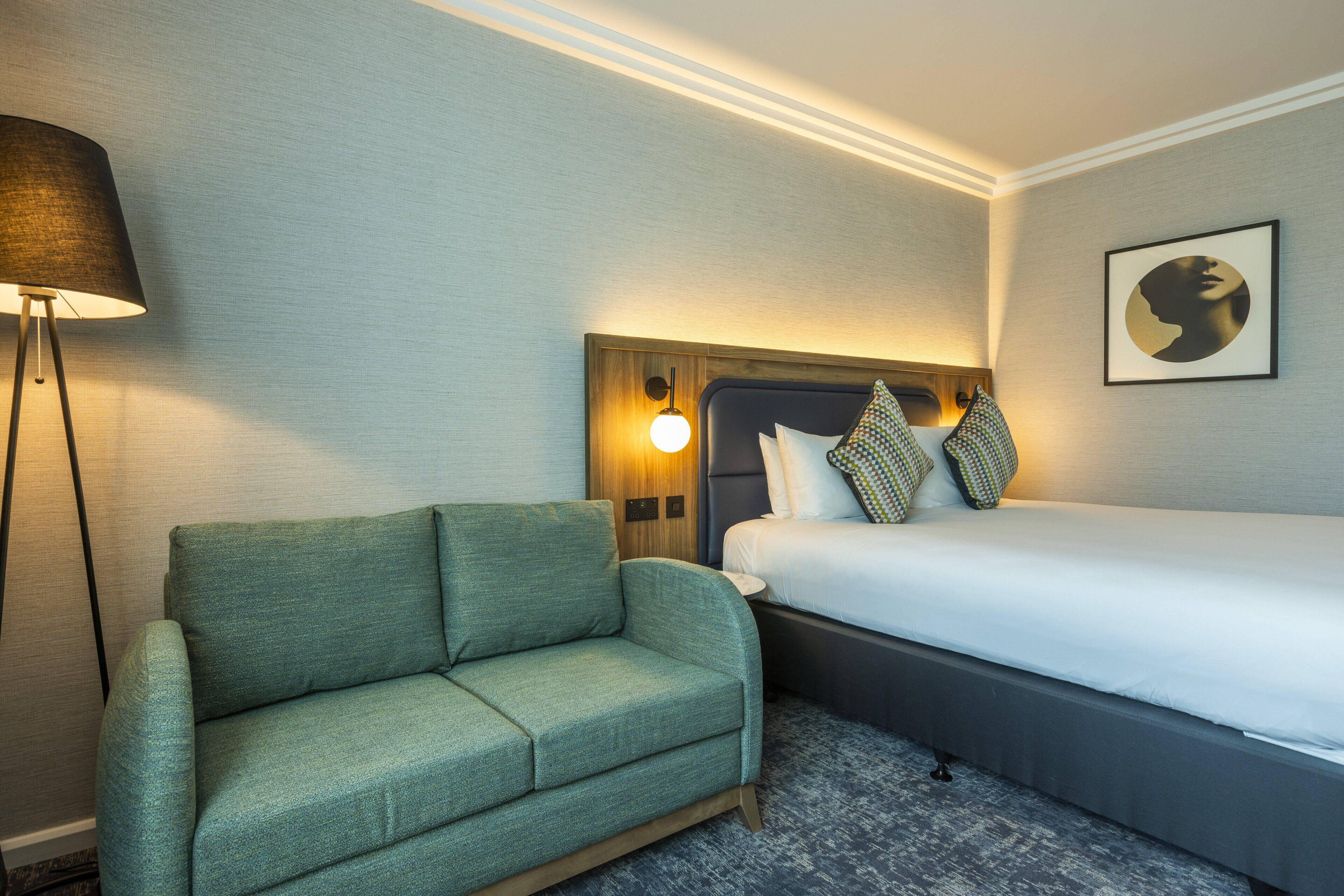 Une chambre d’hôtel avec un canapé vert, un lit avec une literie blanche, une lampe fixée au mur et un tableau encadré accroché au mur.