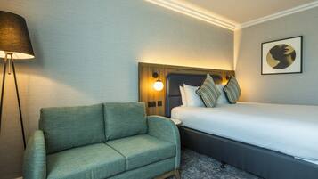 A hotel room with a green sofa, a bed with white bedding, a wall-mounted lamp, and a framed picture on the wall.