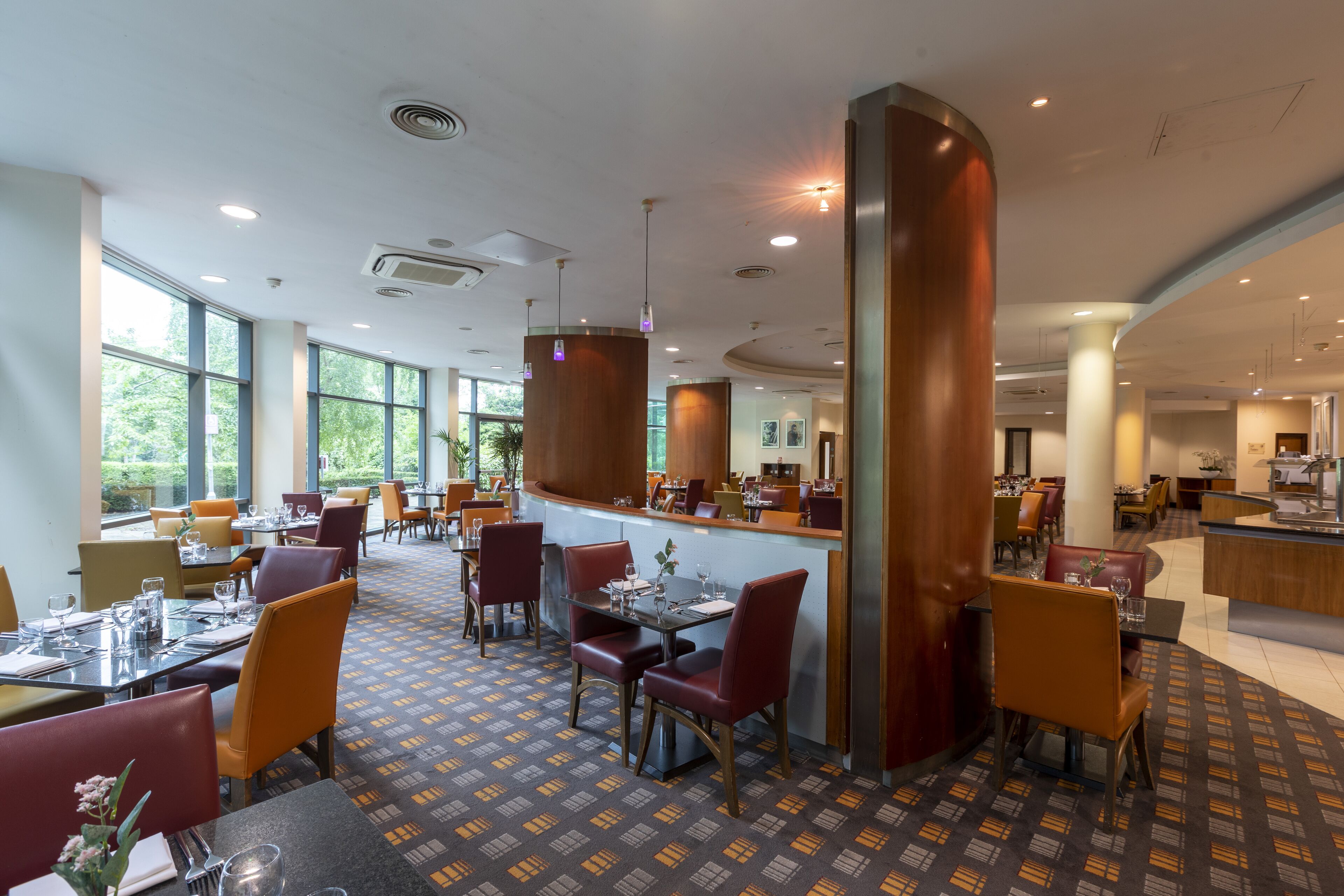 A spacious restaurant with high ceilings, large windows, and a patterned floor. Tables are set with chairs, and there are multiple dining areas visible.