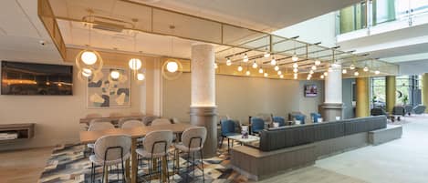 A modern hotel lobby with a central seating area, a high ceiling with hanging lights, and a large window providing natural light.