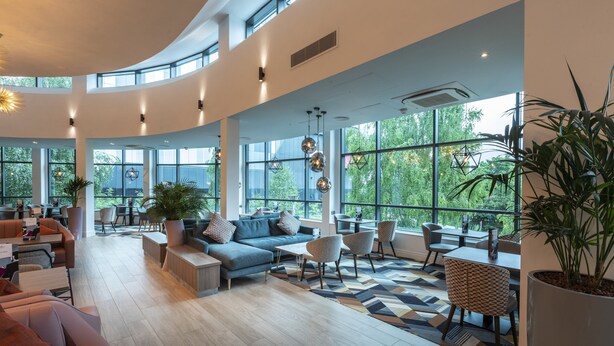 A spacious, well-lit lounge area with a curved ceiling, large windows offering views of greenery, and a variety of seating options including sofas, chairs, and a dining table. - Crowne Plaza Birmingham NEC by IHG (Birmingham)