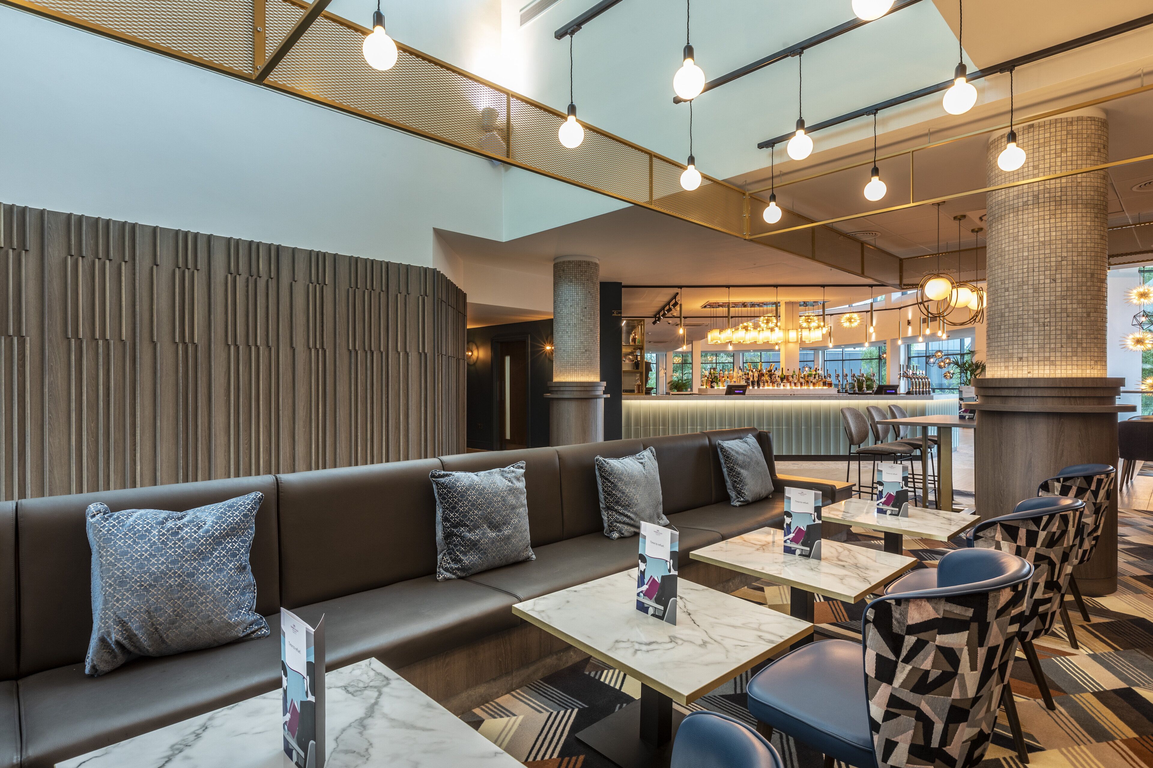 Ein modernes Restaurant mit Barbereich, Sitzgelegenheiten mit gemusterten Stühlen und einer hohen Decke mit hängenden Lampen.