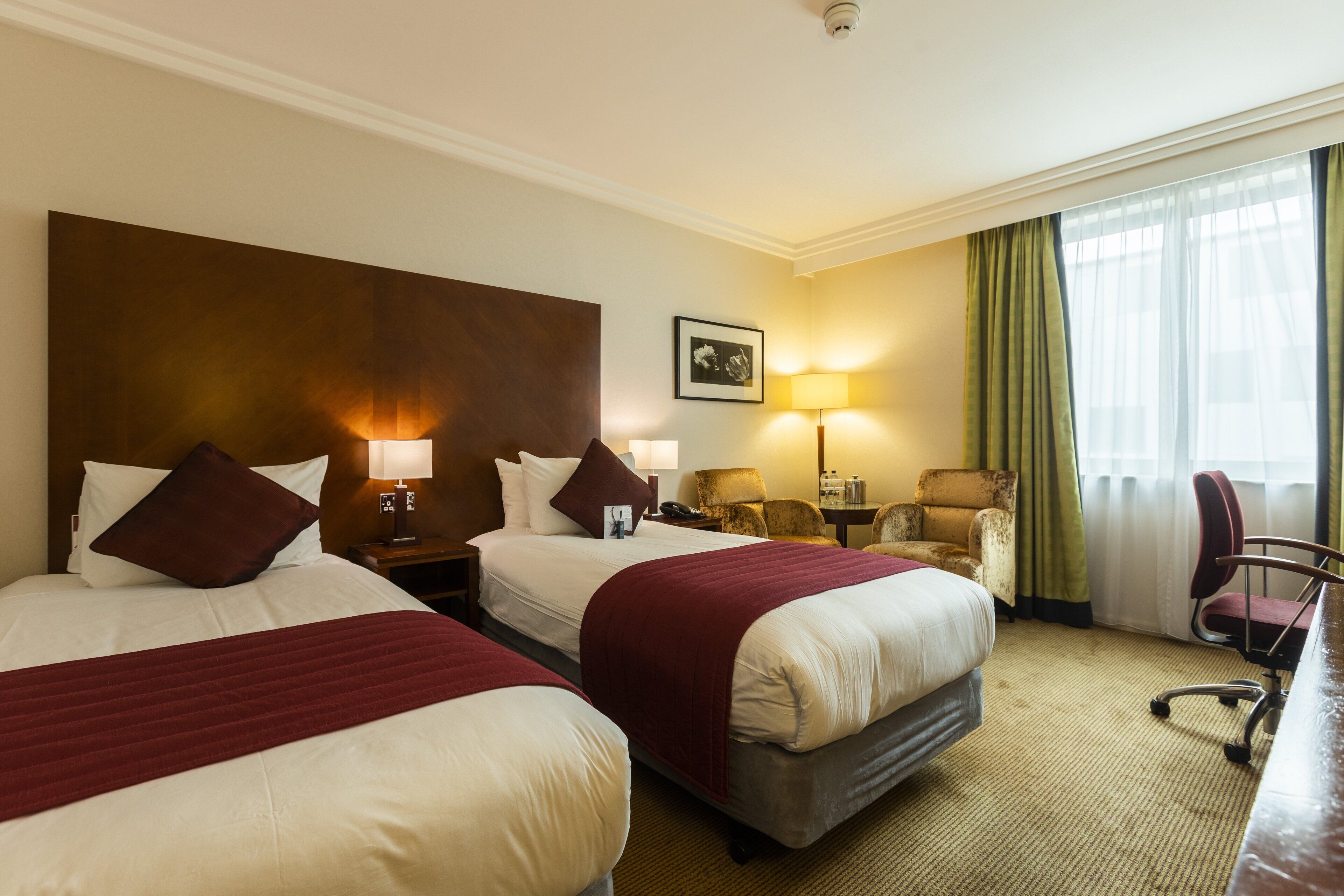 A hotel room with two beds, a desk, a chair, a lamp, and a picture on the wall.