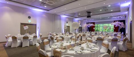 A banquet hall with round tables set for dining, white chairs with gold sashes, and a large decorative centerpiece.