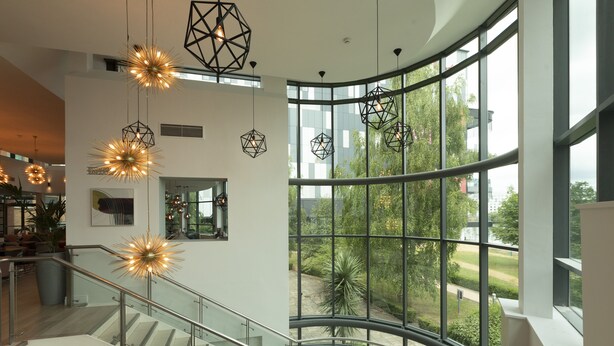 A modern interior with a curved glass railing, geometric pendant lights, and a view of greenery outside. - Crowne Plaza Birmingham NEC by IHG (Birmingham)