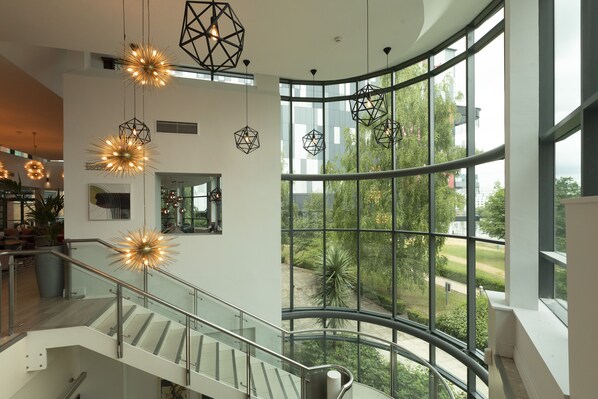 Ein modernes Interieur mit einer gebogenen Glasreling, geometrischen Pendelleuchten und Blick auf Grünflächen.