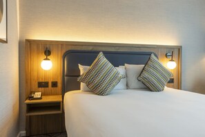 A neatly made bed with two patterned pillows, a bedside table with a phone, and wall-mounted lamps. - Crowne Plaza Birmingham NEC by IHG (Birmingham)