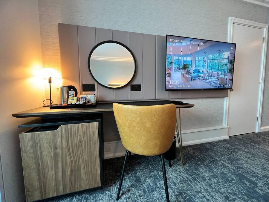 A hotel room with a desk, a round mirror, a television displaying the Crowne Plaza Birmingham NEC, and a yellow chair.