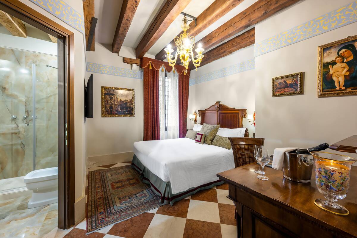 A hotel room with a bed, a chandelier, a painting, a wooden table, and a shower area.