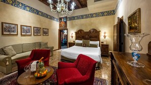 A hotel room with a bed, a red armchair, a sofa, a wooden coffee table with fruit, a chandelier, and framed pictures on the walls. - Hotel Palazzo Priuli (Venice)
