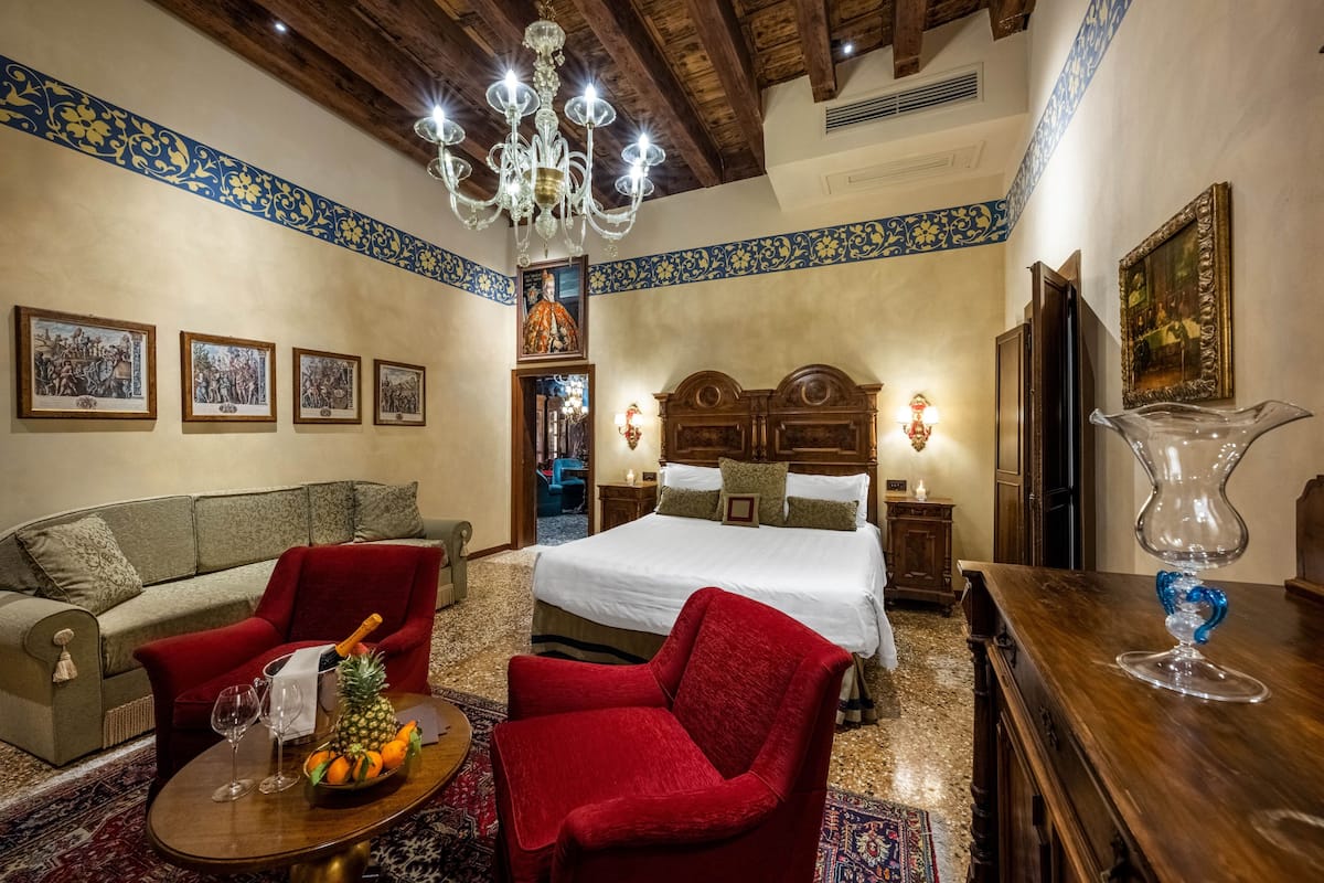 A hotel room with a bed, a red armchair, a sofa, a wooden coffee table with fruit, a chandelier, and framed pictures on the walls.