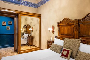 Camera d'albergo con un grande specchio, una testata del letto in legno, un letto con lenzuola bianche e cuscini fantasia, un comodino con una lampada e una parete blu con quadri.