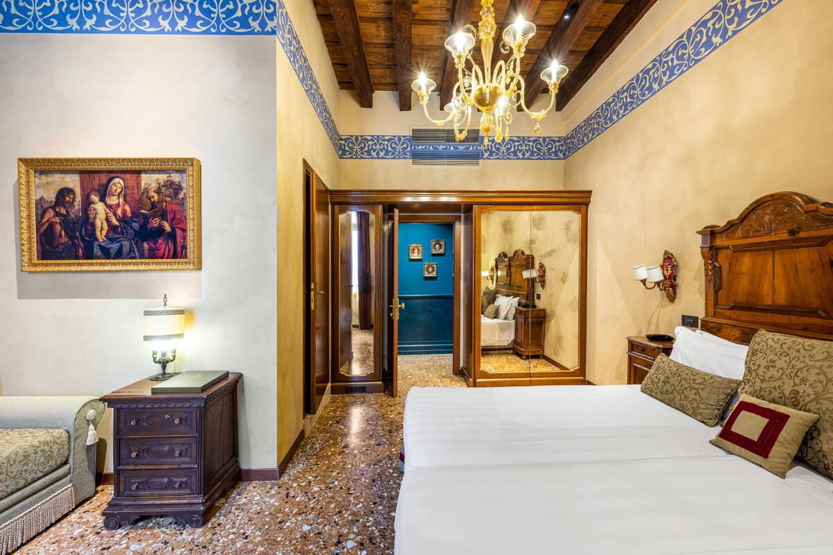 A hotel room with a large bed, a wooden dresser, a nightstand, a chandelier, and a painting on the wall.