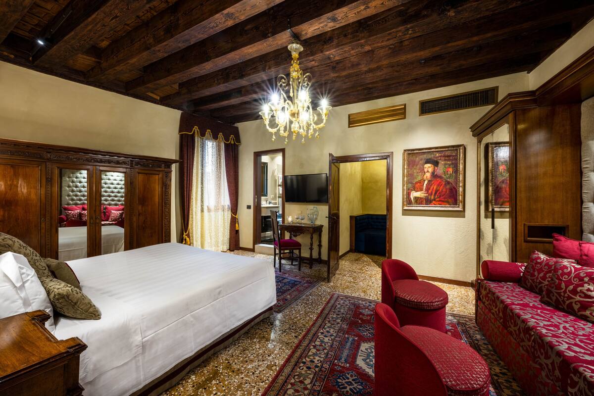 A spacious room with a large bed, a red sofa, a wooden wardrobe, and a chandelier.